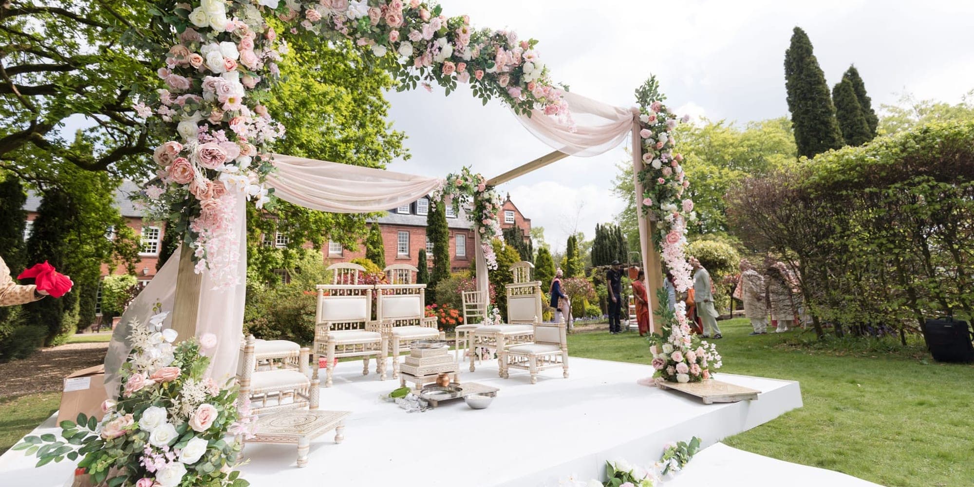 Pink and white floral mandap arch on a white stage in a garden with ceremony seating