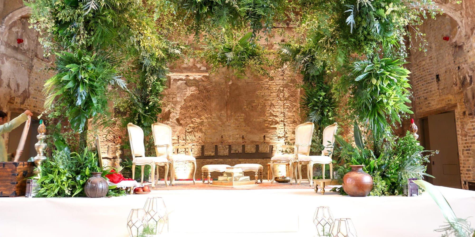 Green foliage frame mandap with ivory chairs against a brick wall