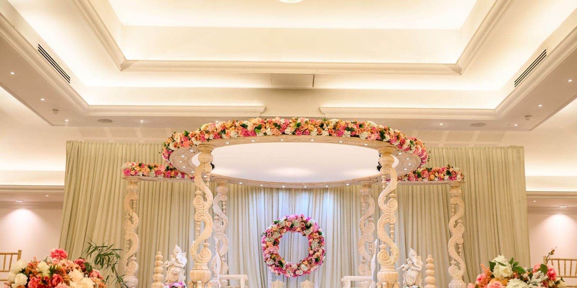 Floral round mandap with white drapes on a stage in a bright hotel ballroom