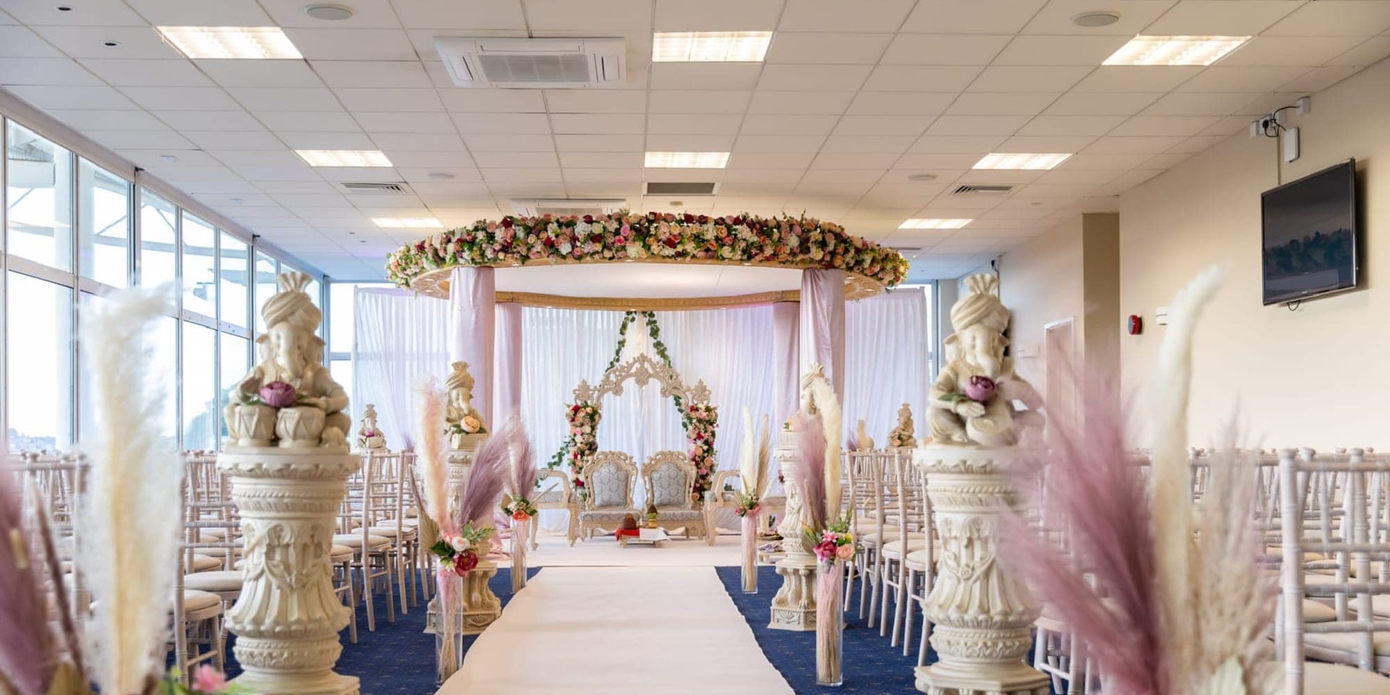 Hall with a white aisle between pedestals leading to a floral canopy mandap