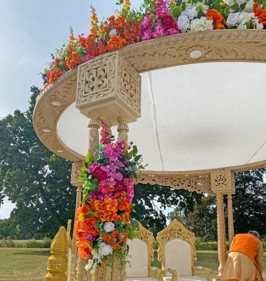 Carved cream round canopy mandap with orange and magenta flowers on a sunny lawn