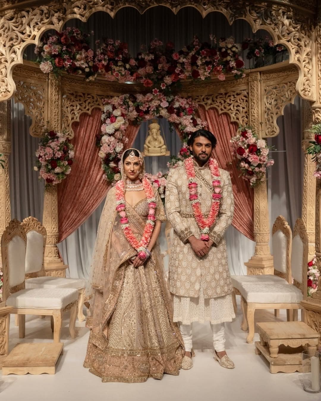 Couple in cream outfits standing before a carved pale wood mandap with terracotta velvet drapes