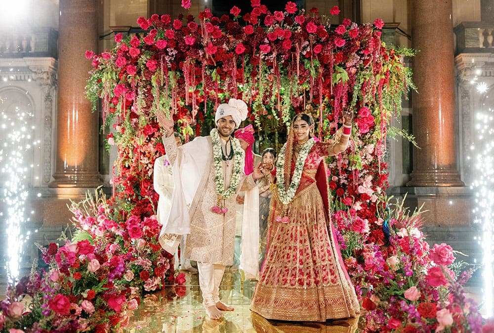 Couple smiling beneath a hanging crimson and magenta floral frame between stone columns and sparklers