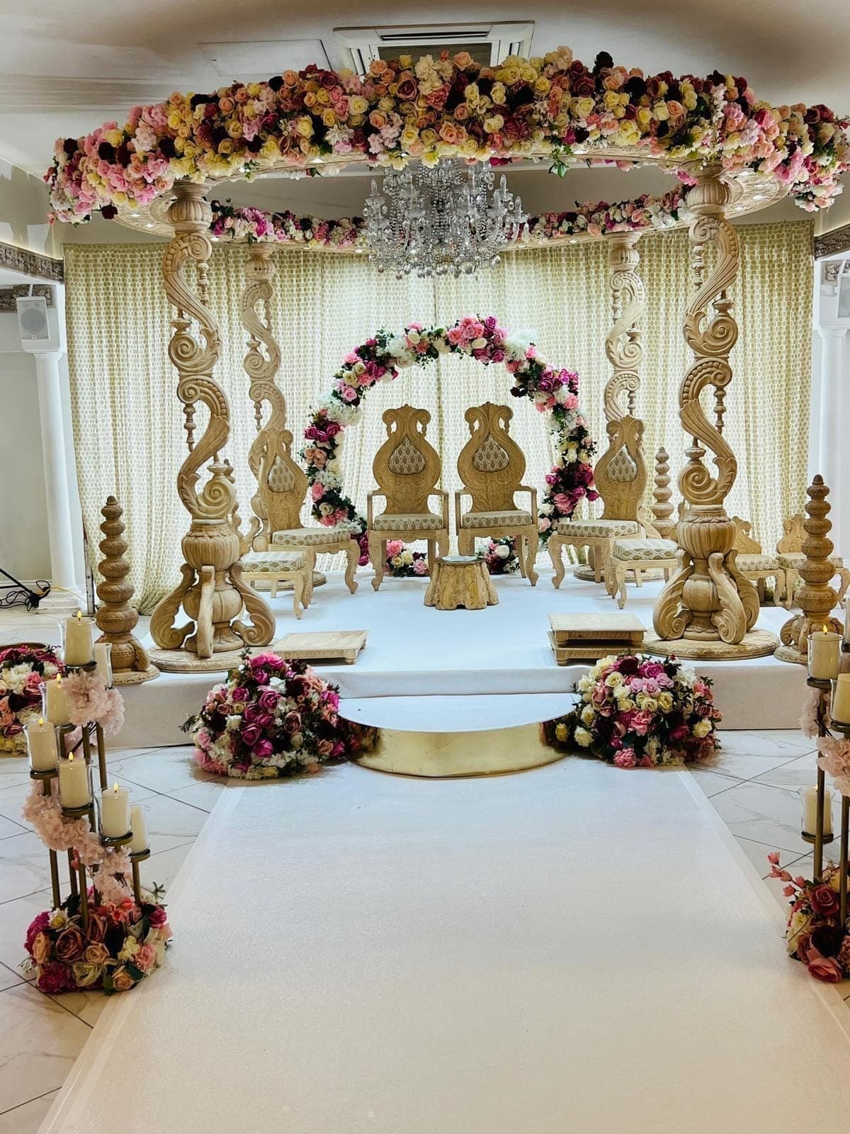 Carved spiral pillar mandap with floral roof and hoop under a crystal light fitting