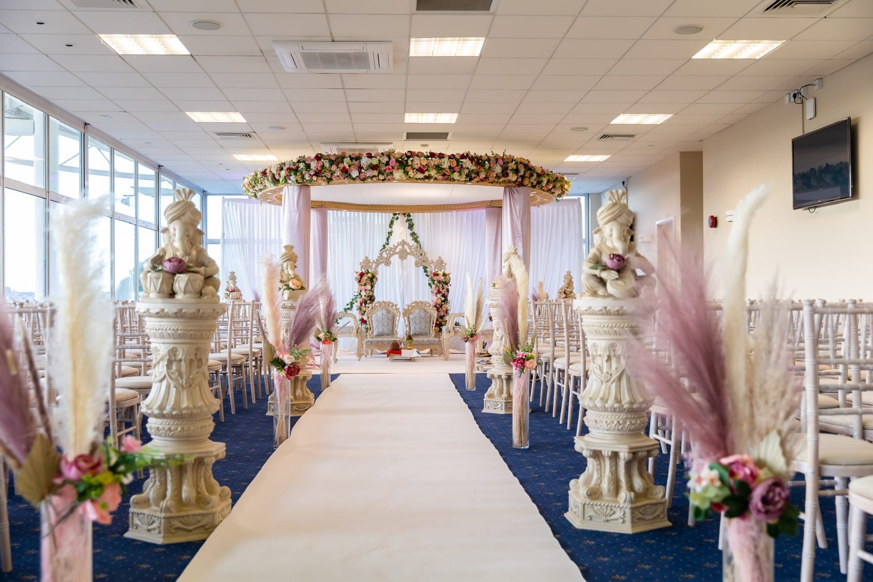 Oval floral mandap at the end of an aisle lined with pampas grass and Ganesh plinths