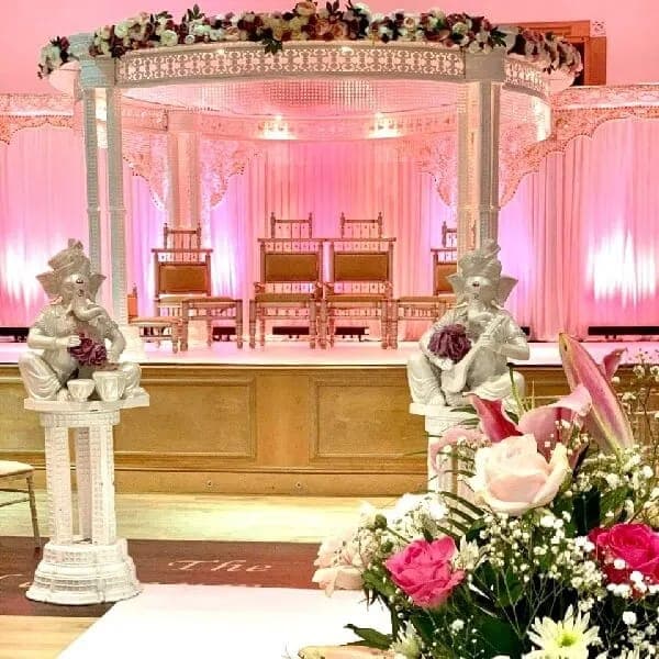 White carved round mandap with pastel flowers and pink lit drapes on a stage