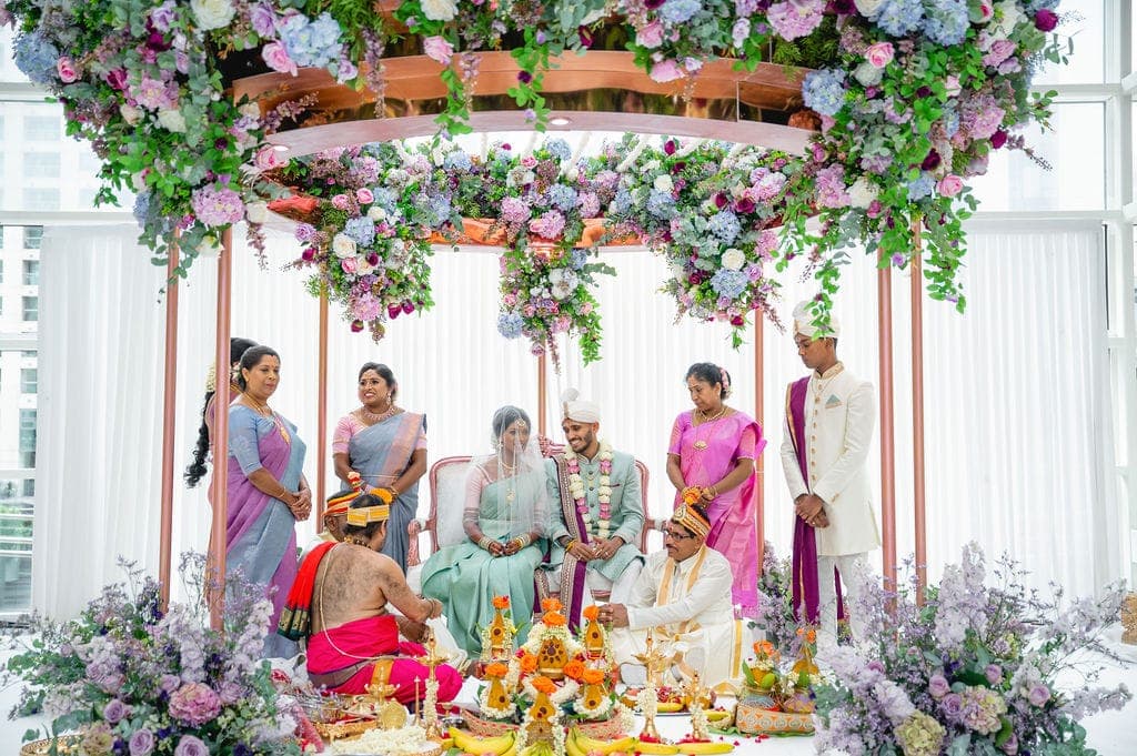 Round rose gold canopy with lilac and blue flowers above a family ceremony beside tall windows