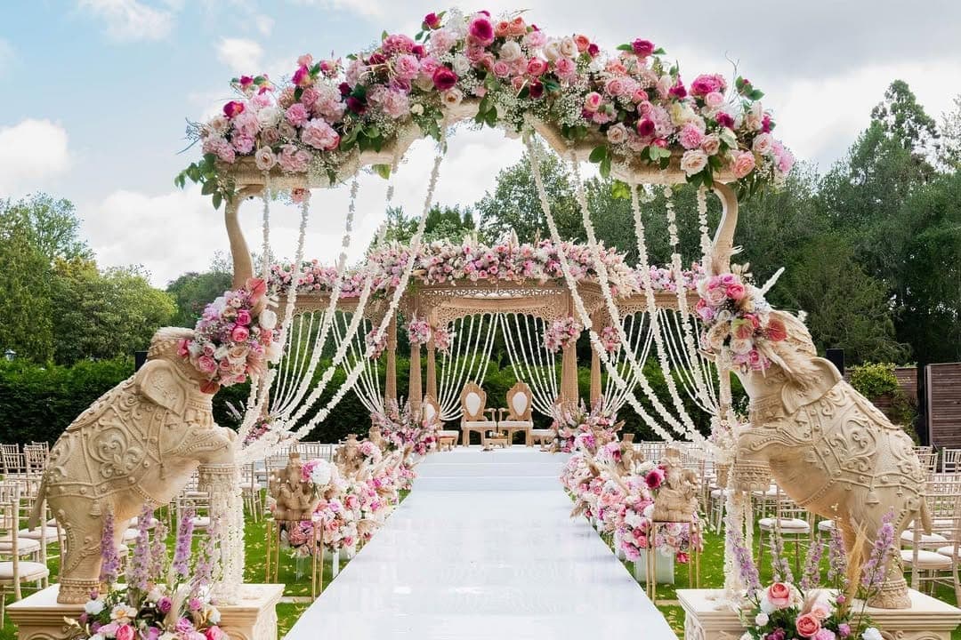 Two carved elephants holding a flowered arch with garlands over a white aisle, pavilion beyond