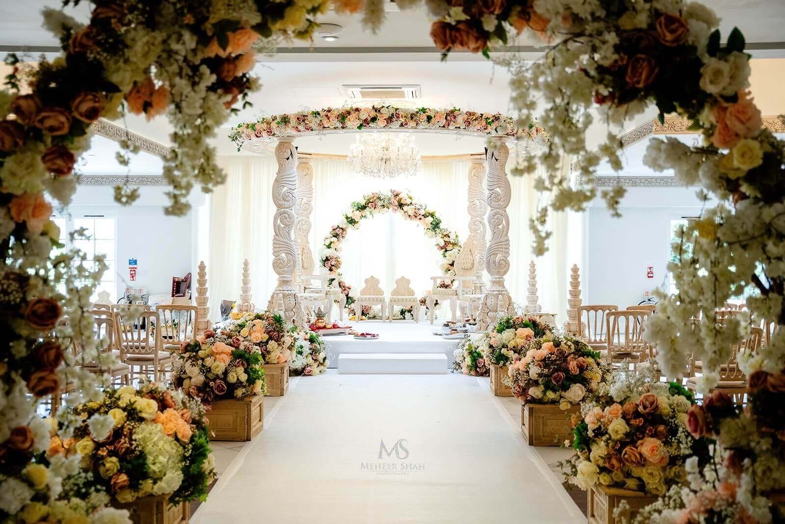 Floral arches along a white aisle leading to a carved mandap and floral ring