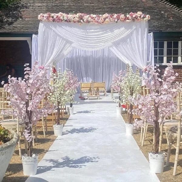 White chiffon frame with pink floral garland and blossom trees in pots lining a white aisle