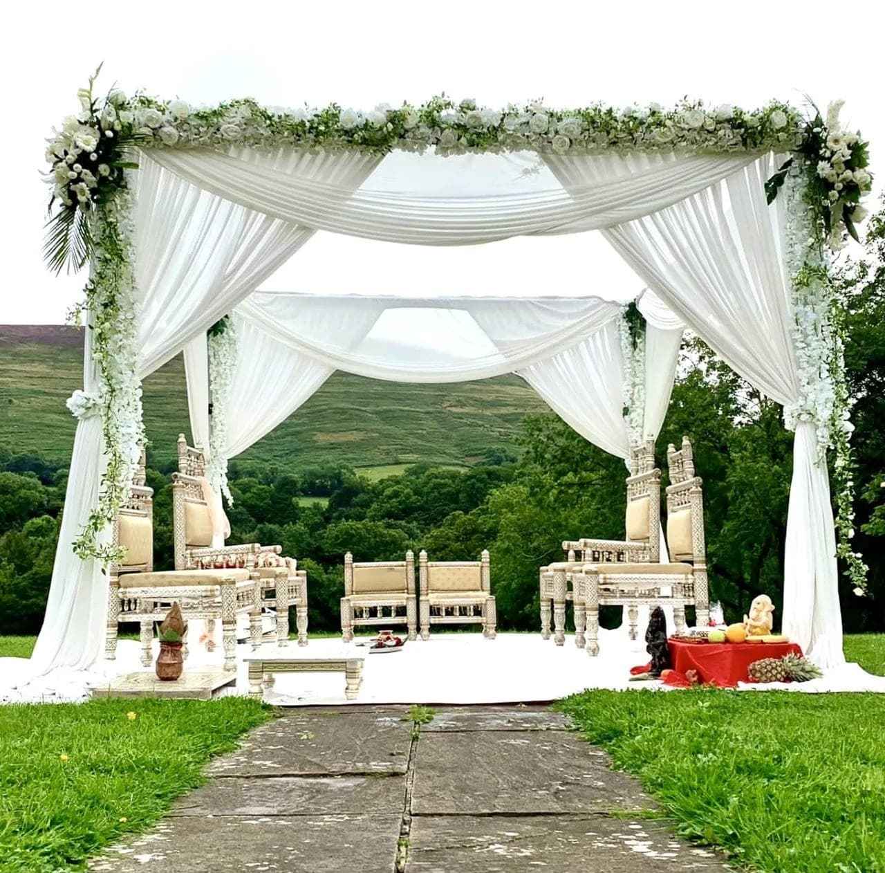 White chiffon draped mandap with white rose garland on a lawn before green hills