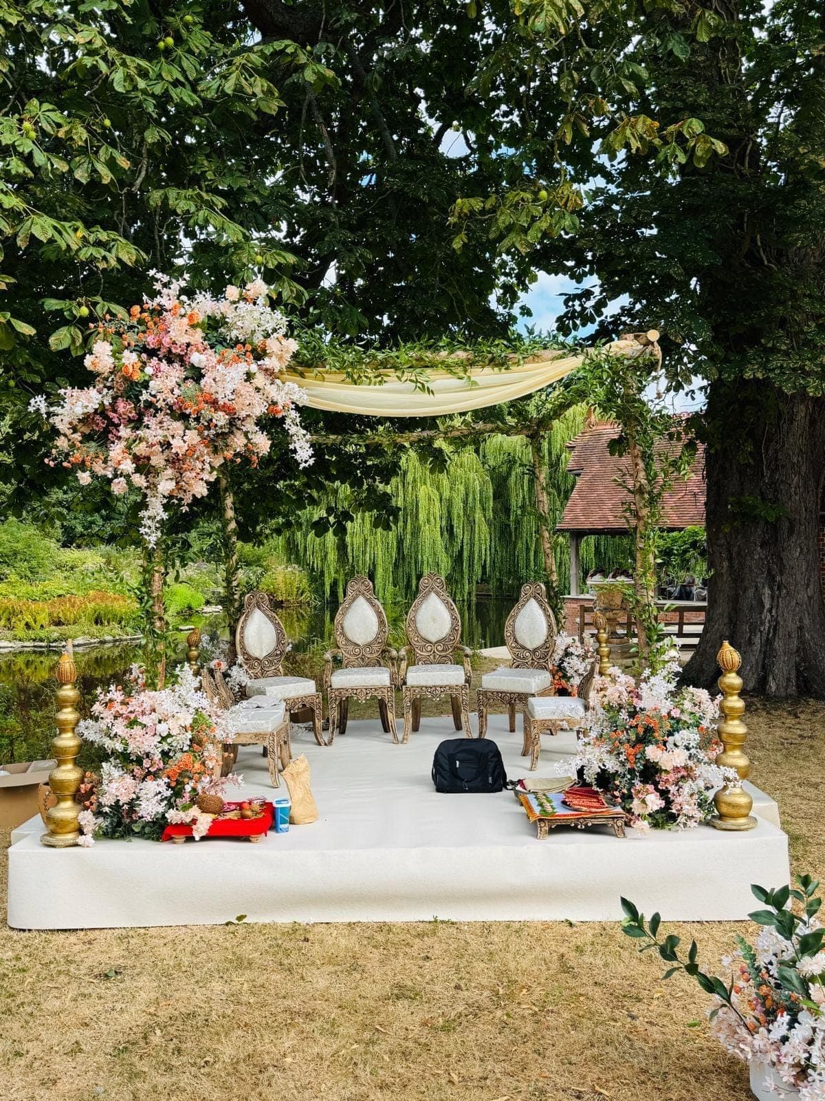 Lawn mandap with cream canopy swag, tall blush florals and ivory chairs under trees