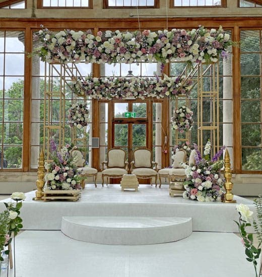 Gold frame mandap with lilac and white flowers inside a bright glasshouse