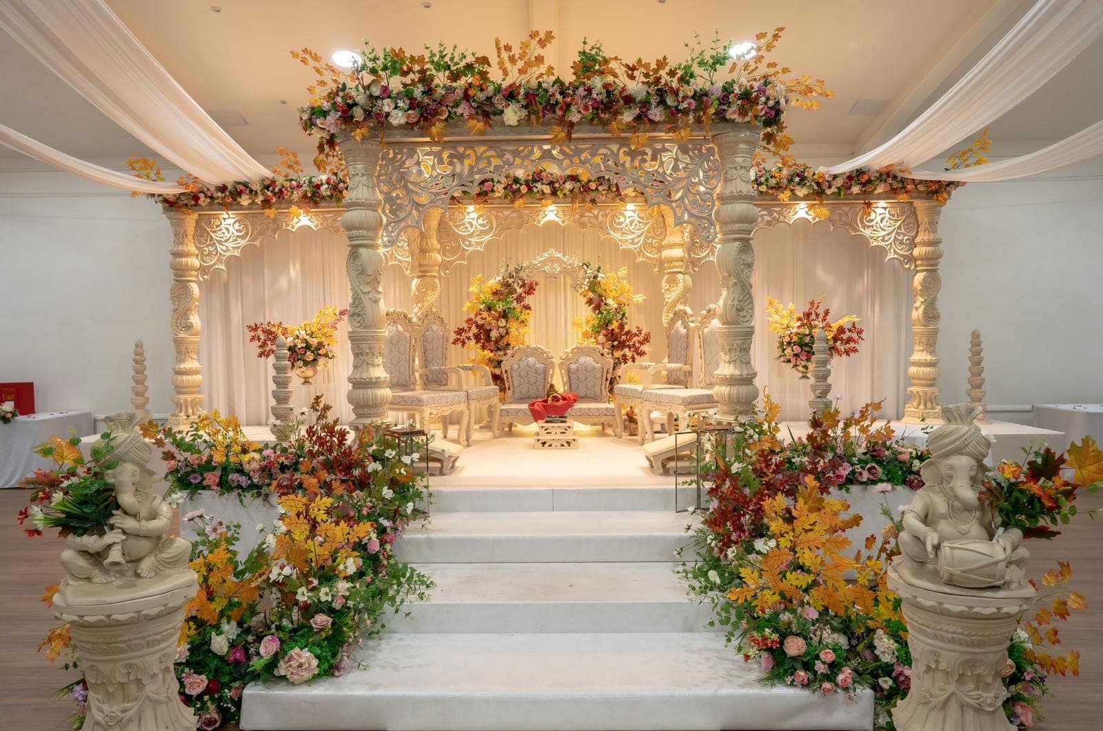 Cream carved mandap with autumn leaves and warm lights above floral steps