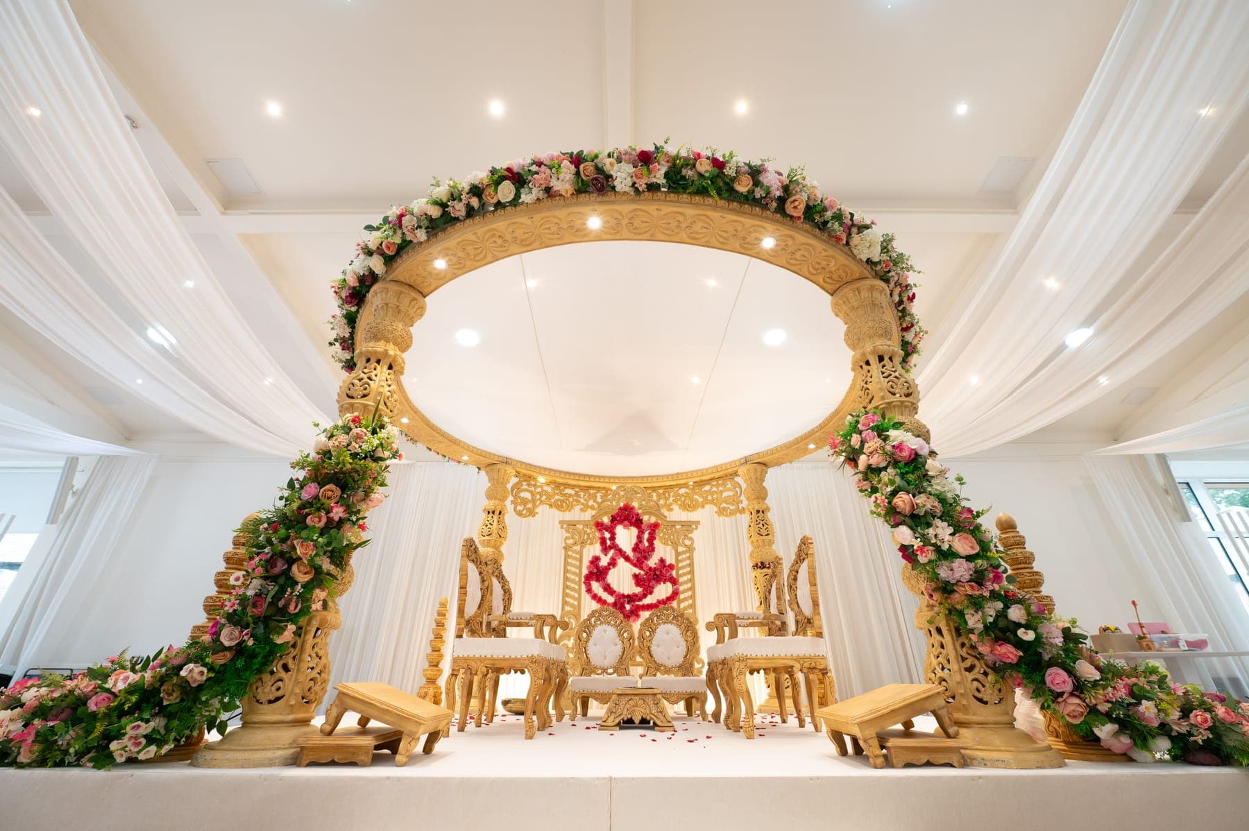 Gold round mandap with trailing pink garlands, red rose Ganesh backdrop and white drapes