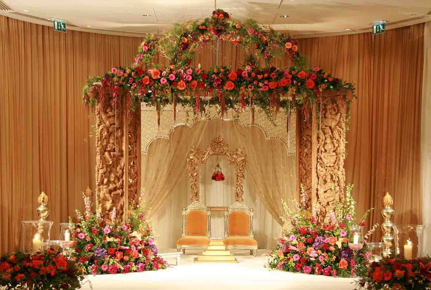 Carved wooden mandap panels with red roses above a gilt frame and orange chairs