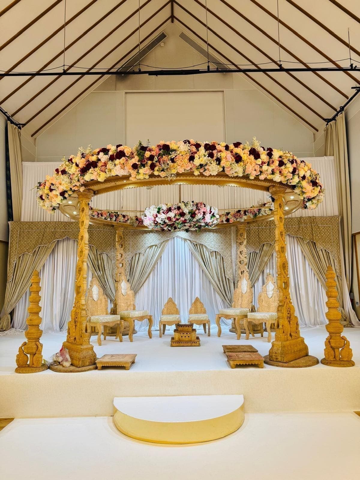 Gold post mandap with a wide floral roof, white stage and gold half-moon step