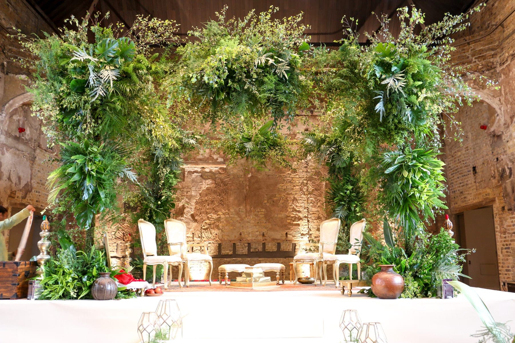 Leafy fern and monstera arch against an exposed brick wall with cream chairs