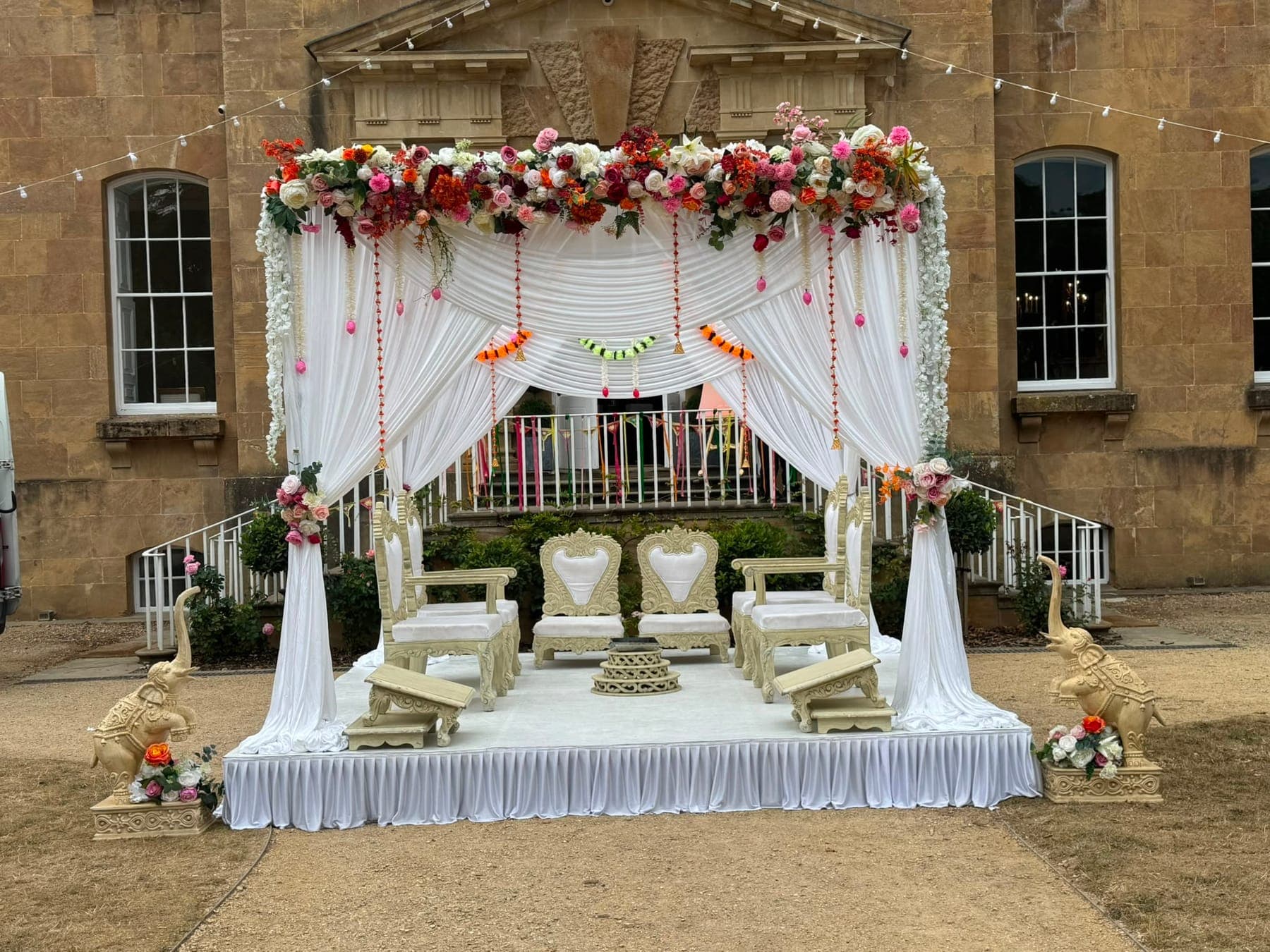 White draped canopy with a pink and orange floral top on a skirted stage in front of a manor house