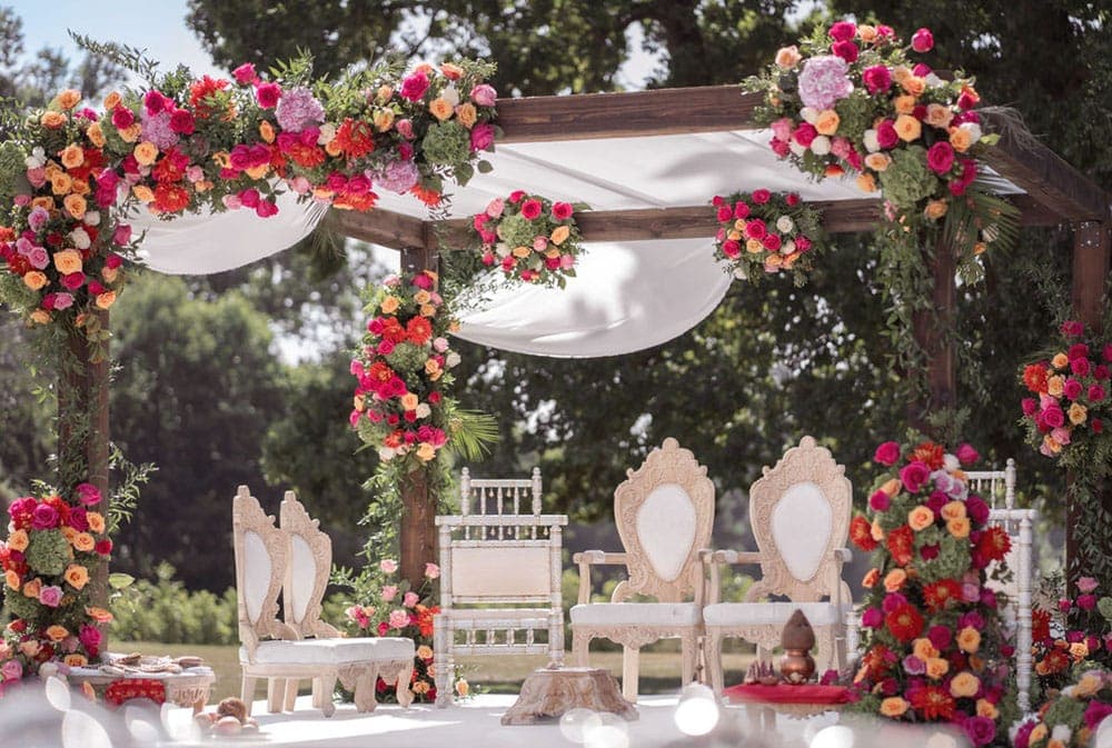 Wooden four-post mandap draped in white with orange, red and magenta flowers and carved ivory chairs