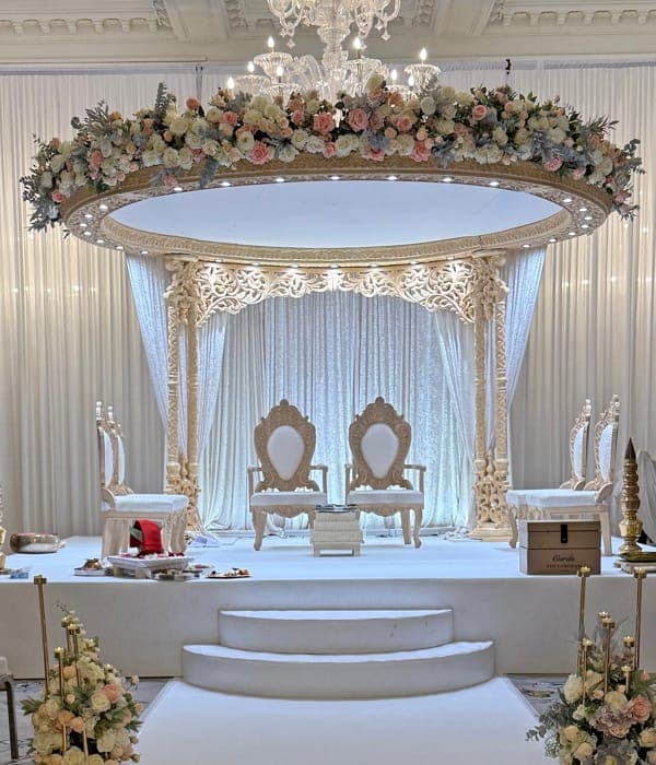 Oval carved mandap with cream floral garland beneath a chandelier