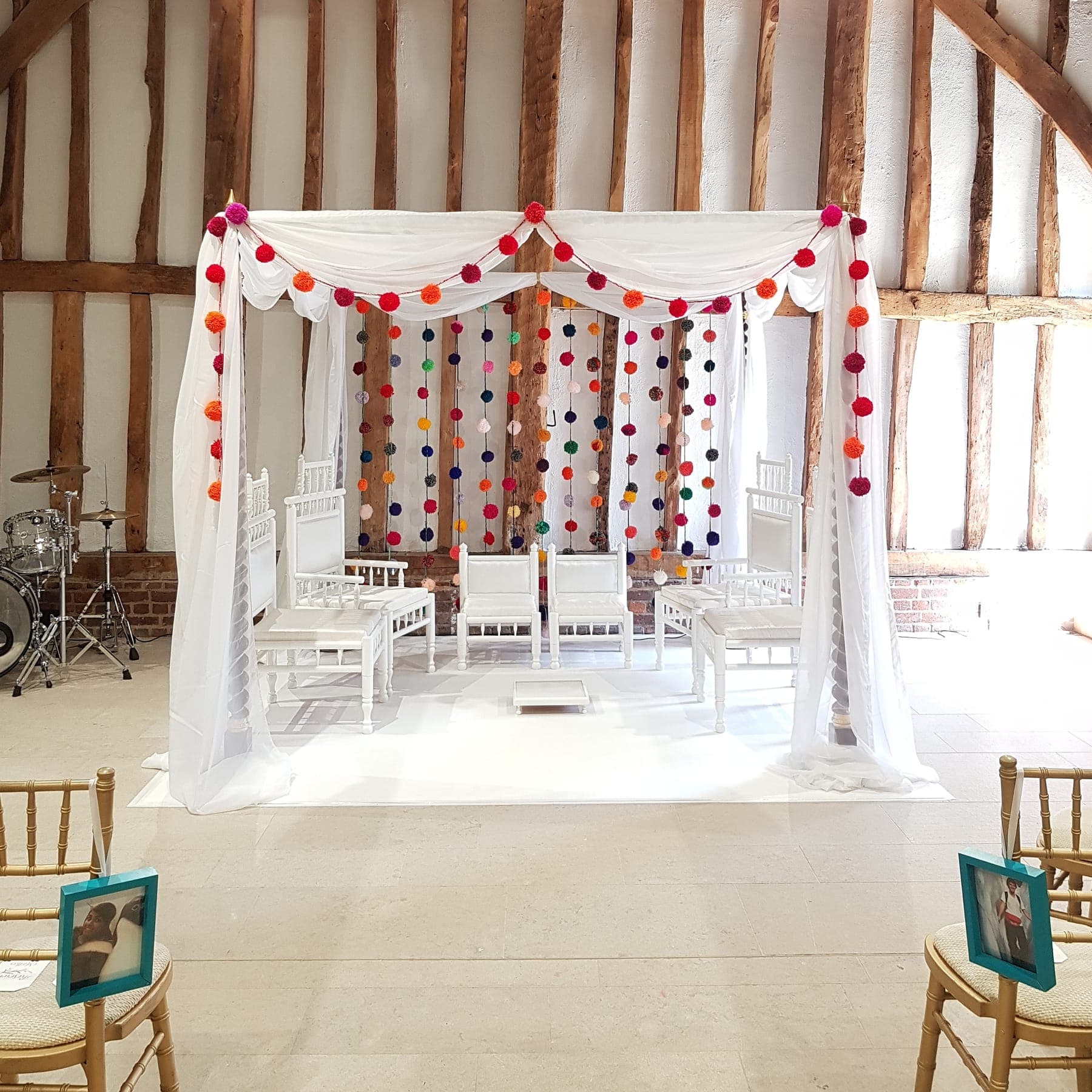 White draped frame with red, orange and pink pom-pom garlands inside a timber-beamed barn