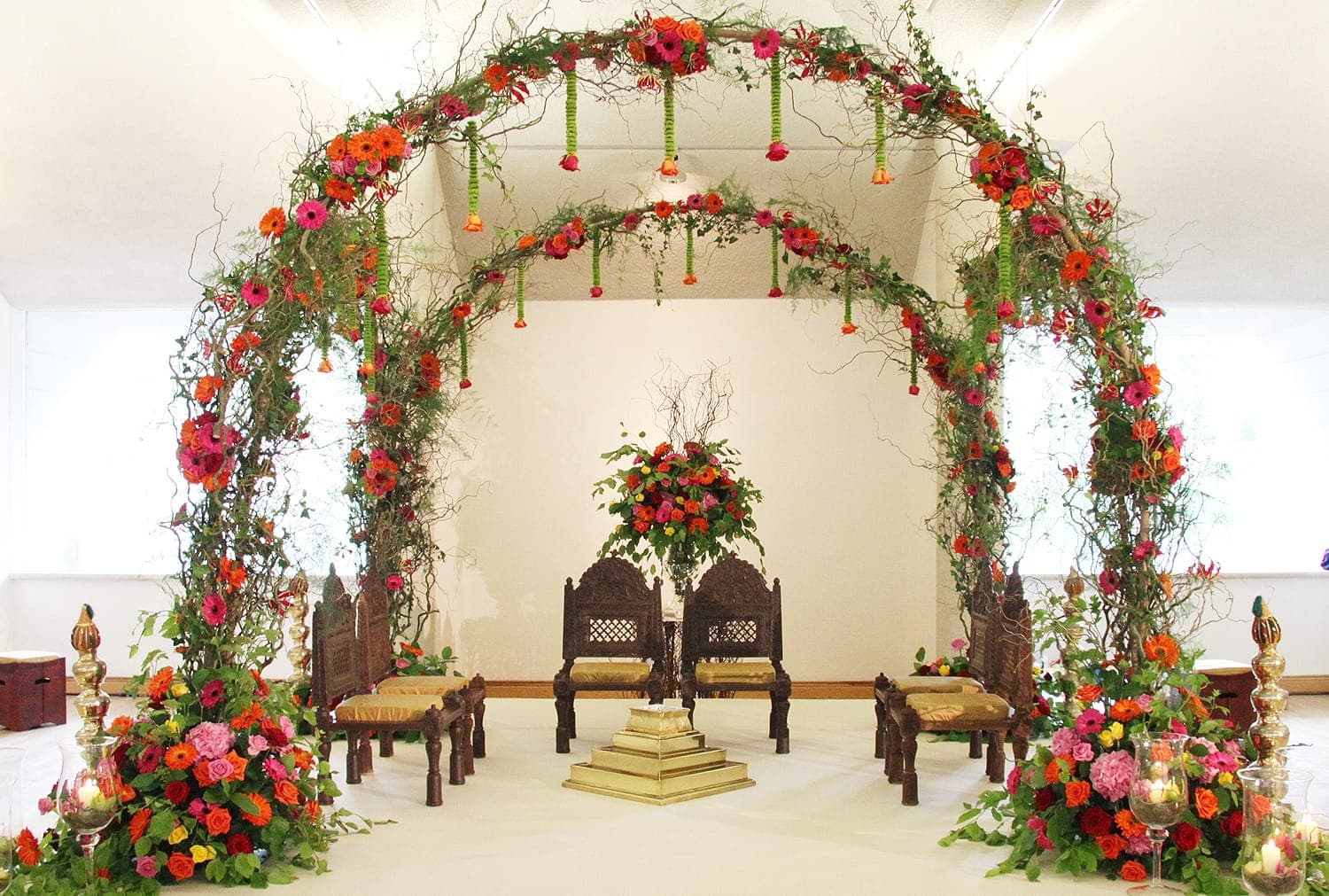 Twig arches with orange and pink blooms over dark carved chairs in a bright room