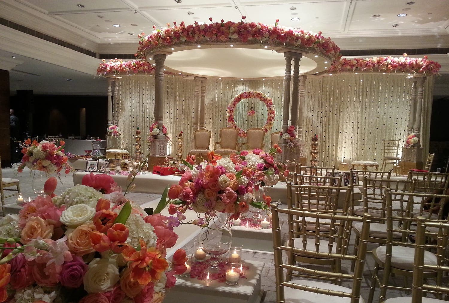 Round floral canopy mandap with coral roses above ivory pillars in a ballroom