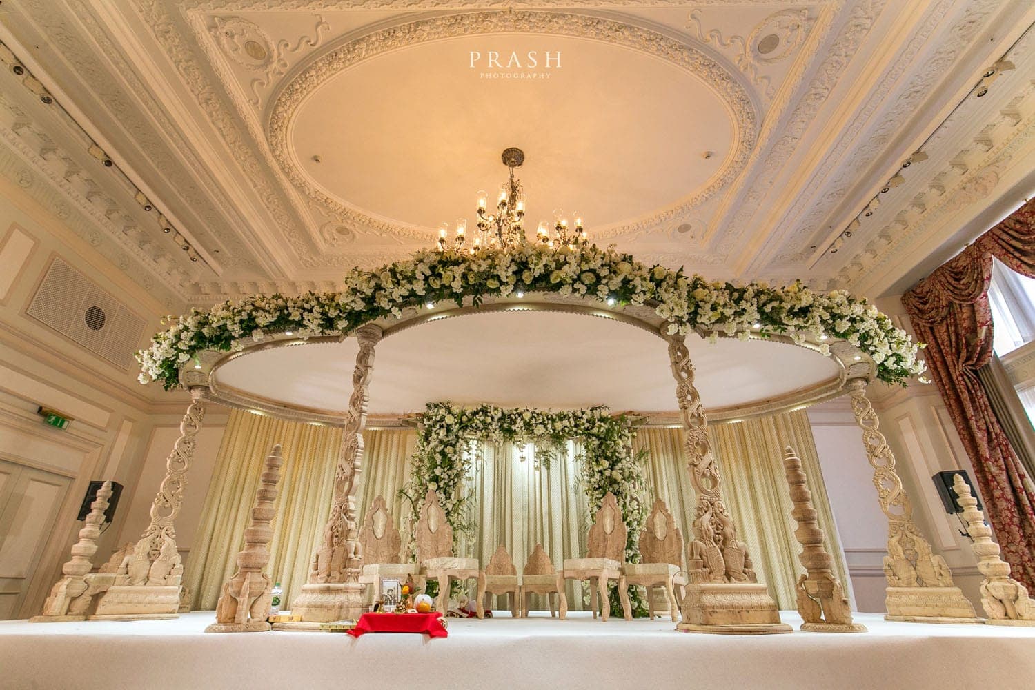 Round carved mandap with white flower garland under an ornate ceiling and chandelier