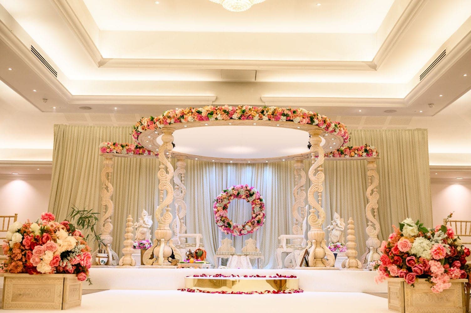 Cream scroll-carved mandap with coral floral crown and round wreath