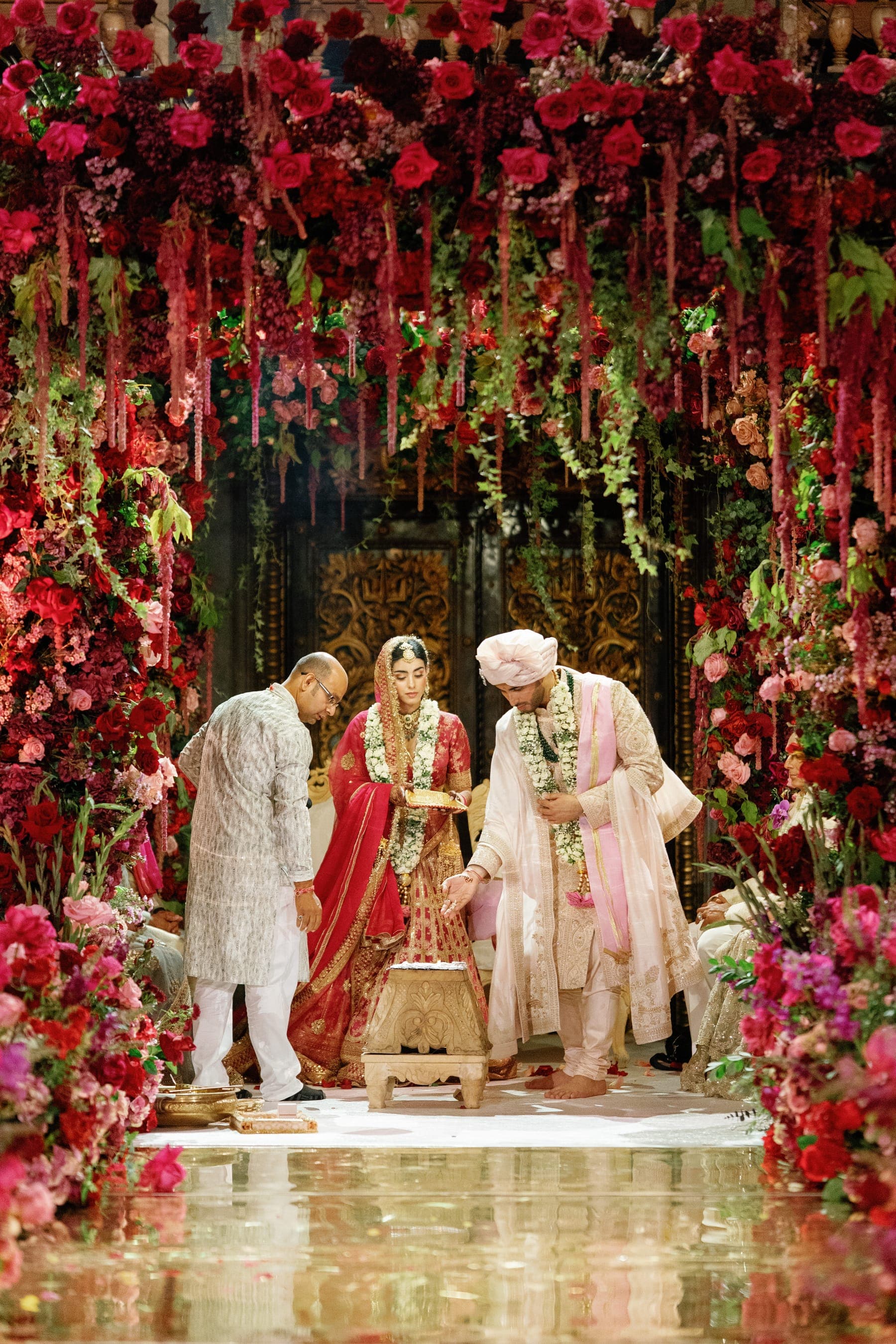 Couple under a dense red rose canopy with trailing amaranthus and a carved gold door