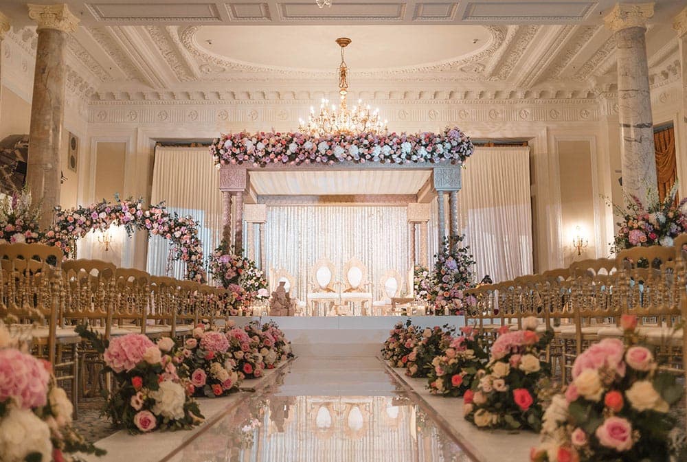Pale pillared mandap with rose garland and ivory drapes at the end of a mirrored aisle