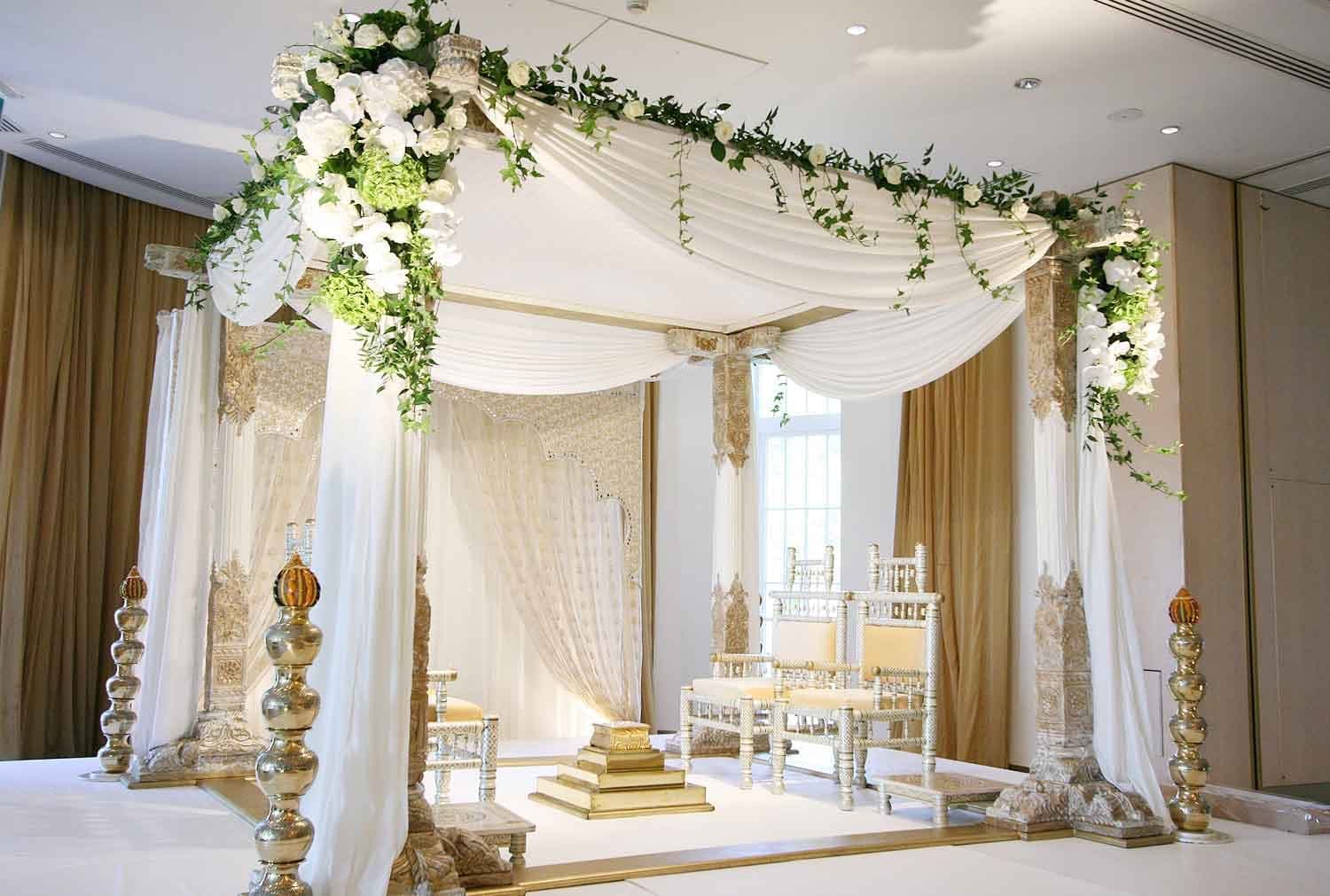 White draped four-post mandap with ivy and white orchids beside brass finials