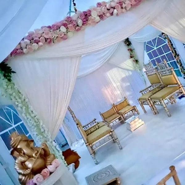 White draped mandap with pink rose garland and gold chairs inside a marquee with arched windows