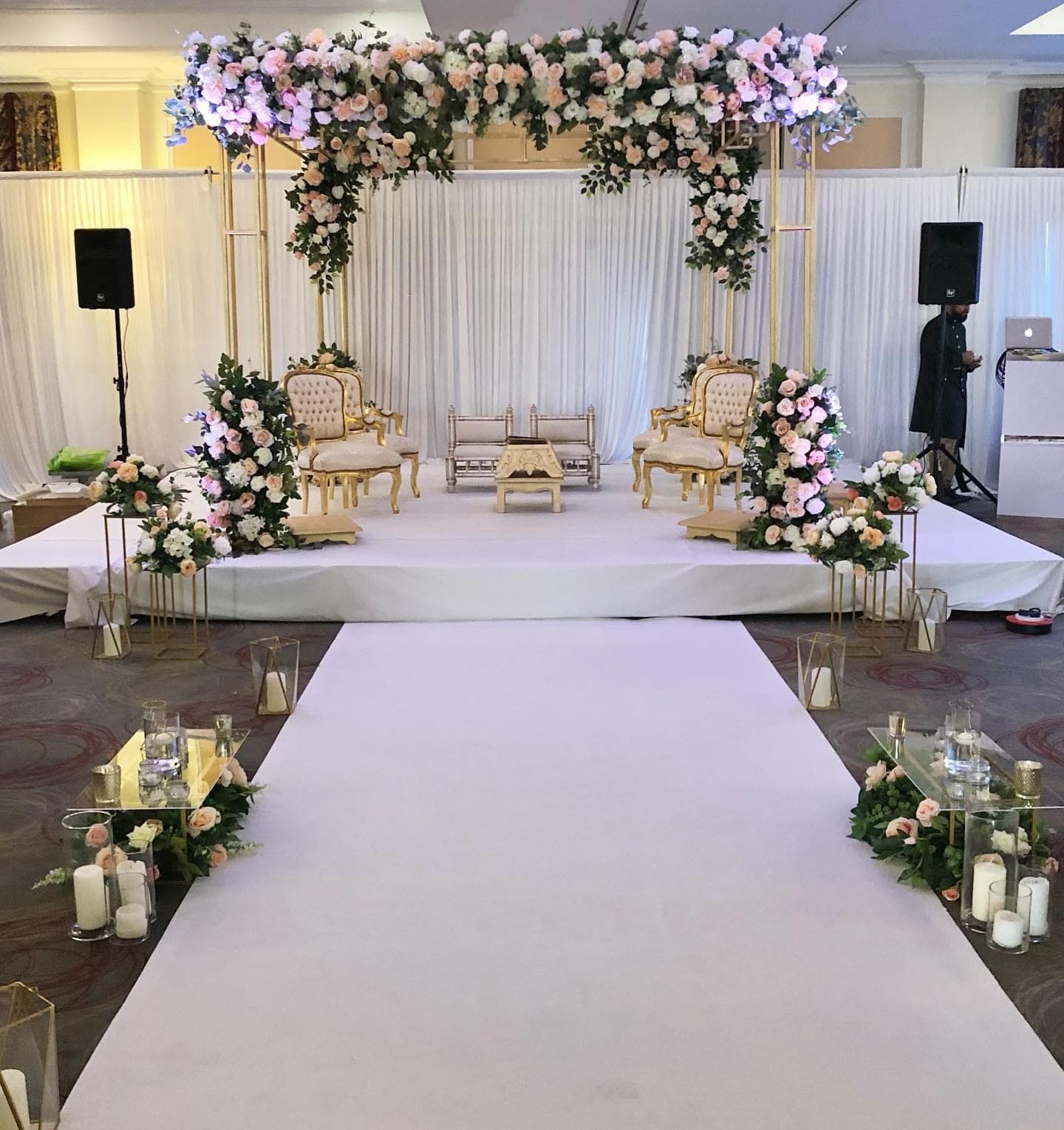 Slim gold frame mandap with peach floral crown and white aisle with lanterns