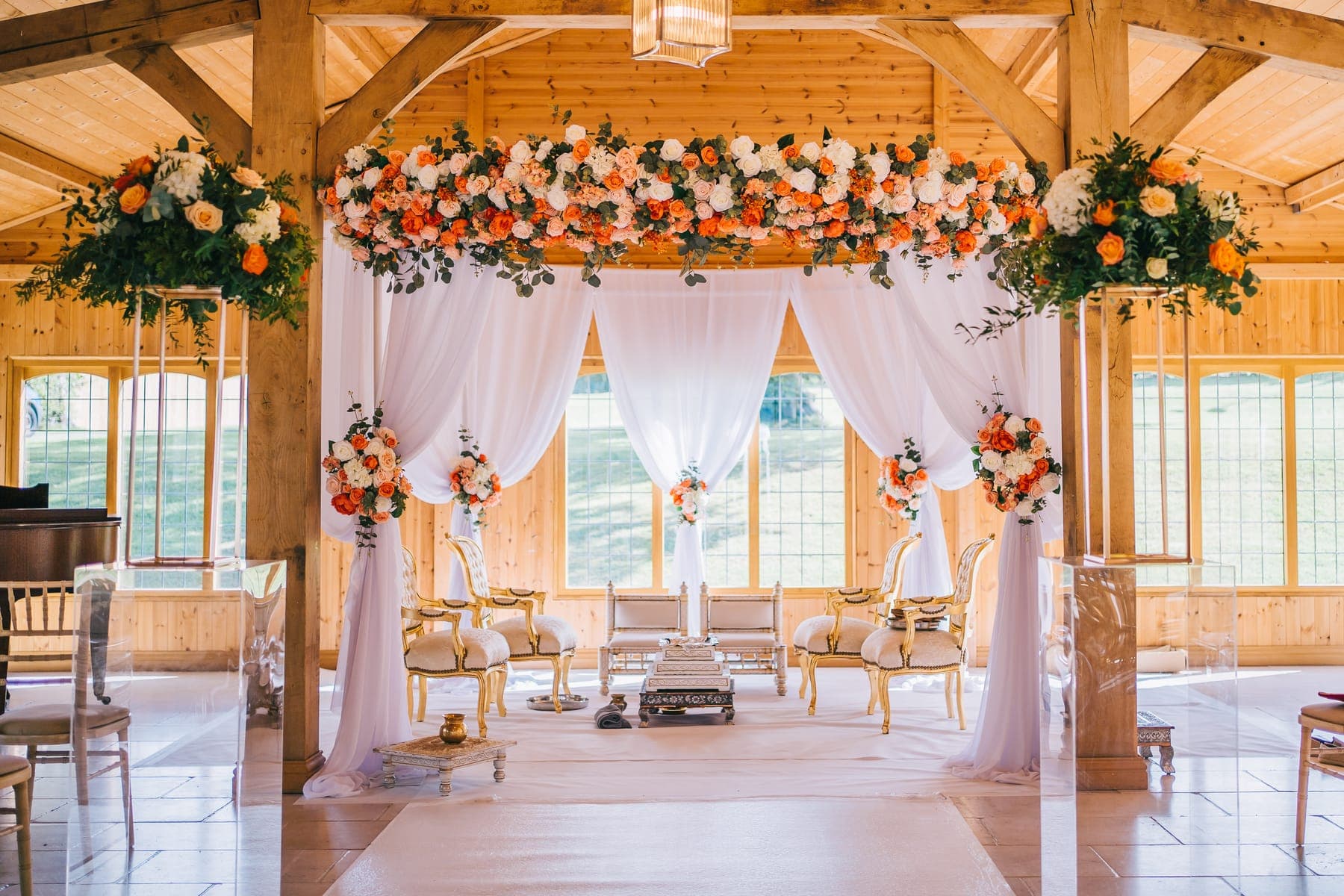 White draped mandap with orange and peach floral crown in an oak hall