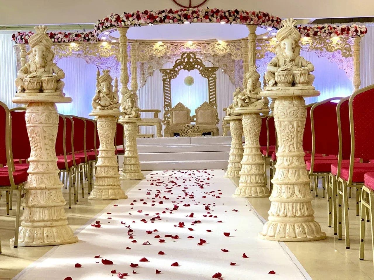 Cream carved pedestals with musician Ganesh figures along a petal-strewn aisle toward a lit carved mandap Cream carved pedestals with musician Ganesh figures along a petal-strewn aisle toward a lit carved mandap