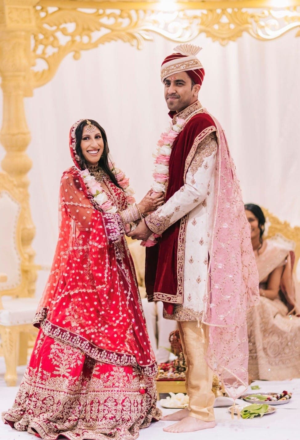 Bride and groom standing beneath cream-gold carved mandap scrollwork and drapes Bride and groom standing beneath cream-gold carved mandap scrollwork and drapes