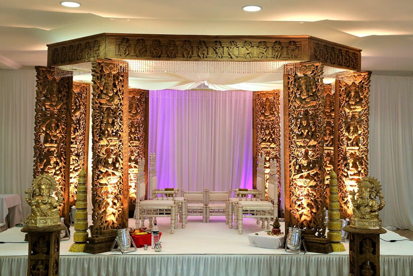 Carved wooden pillar mandap with hanging crystals and ivory drape on a lit stage Carved wooden pillar mandap with hanging crystals and ivory drape on a lit stage