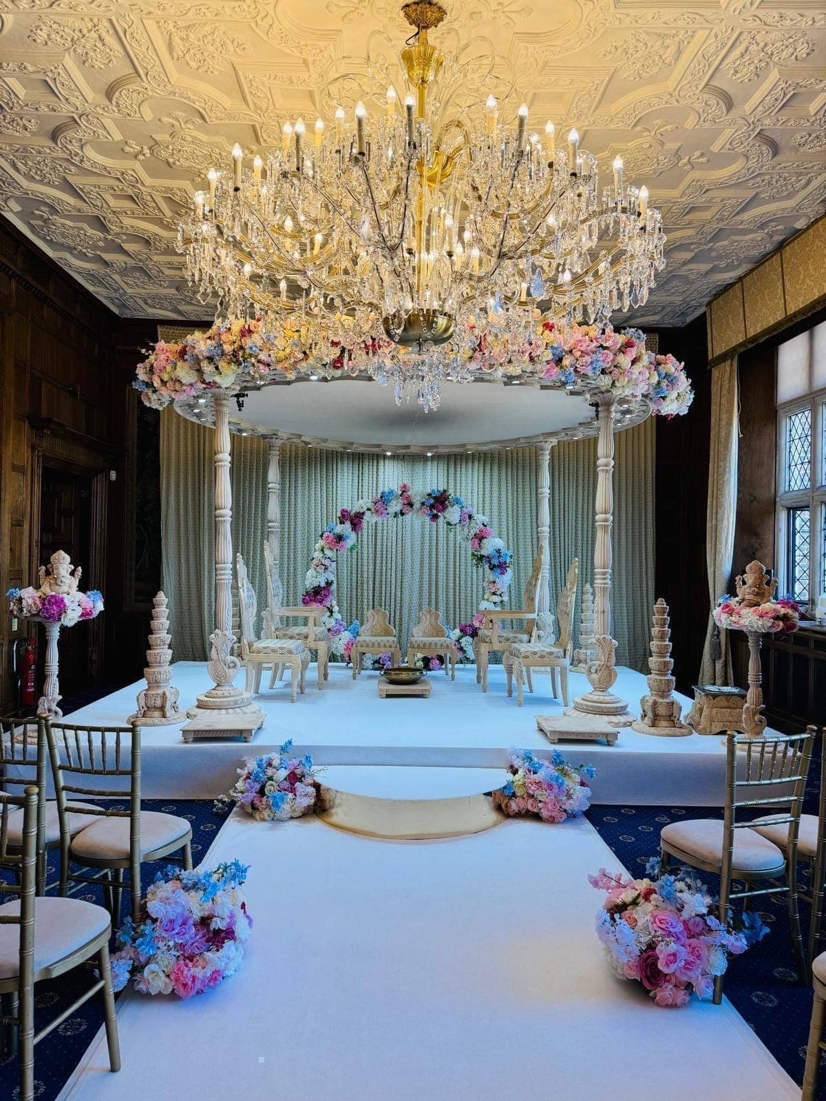 Round floral canopy under a chandelier with a floral hoop and white aisle in a panelled manor room Round floral canopy under a chandelier with a floral hoop and white aisle in a panelled manor room