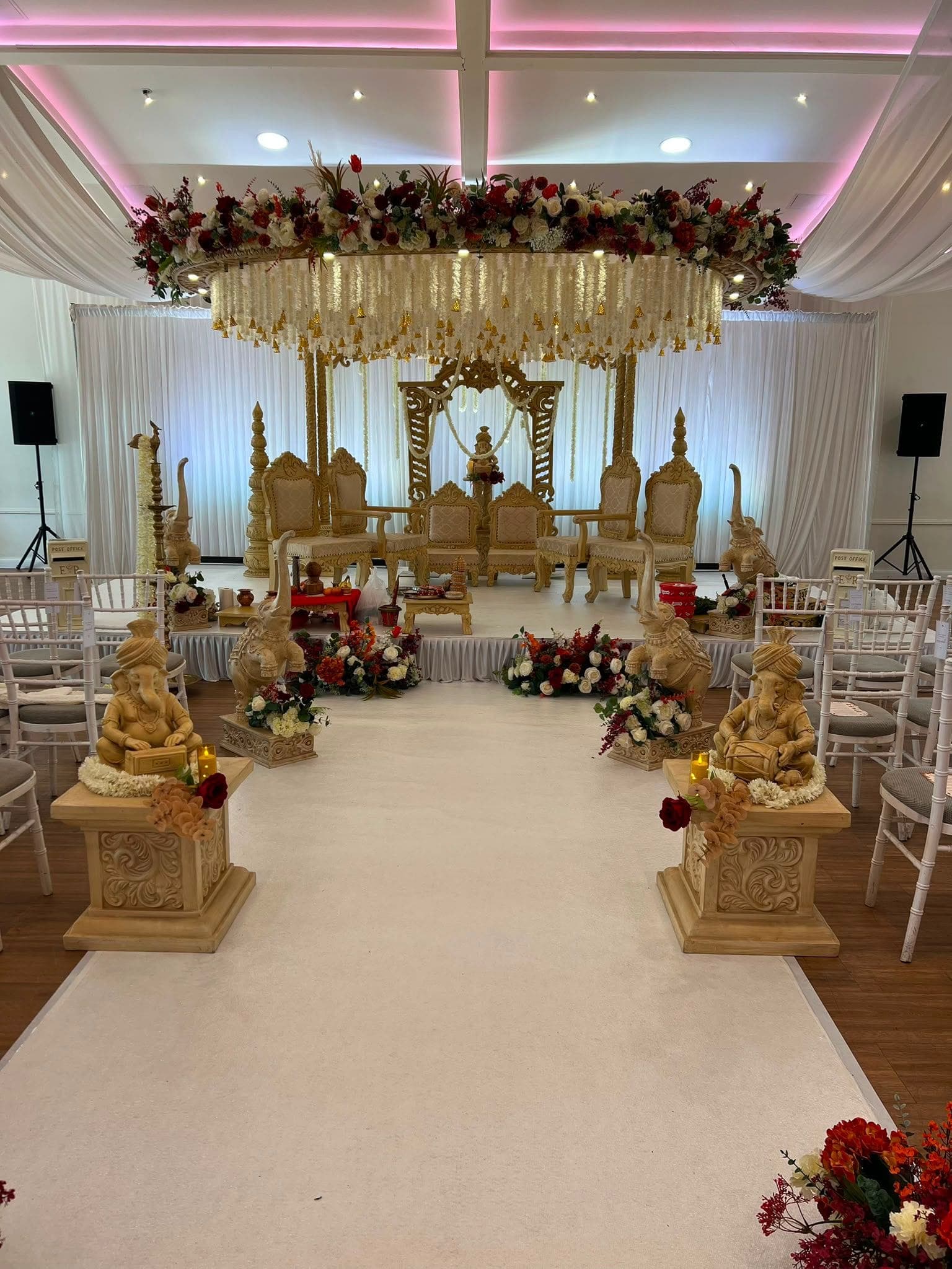 Round floral canopy with hanging cream strands over gold chairs at the end of a pale aisle Round floral canopy with hanging cream strands over gold chairs at the end of a pale aisle