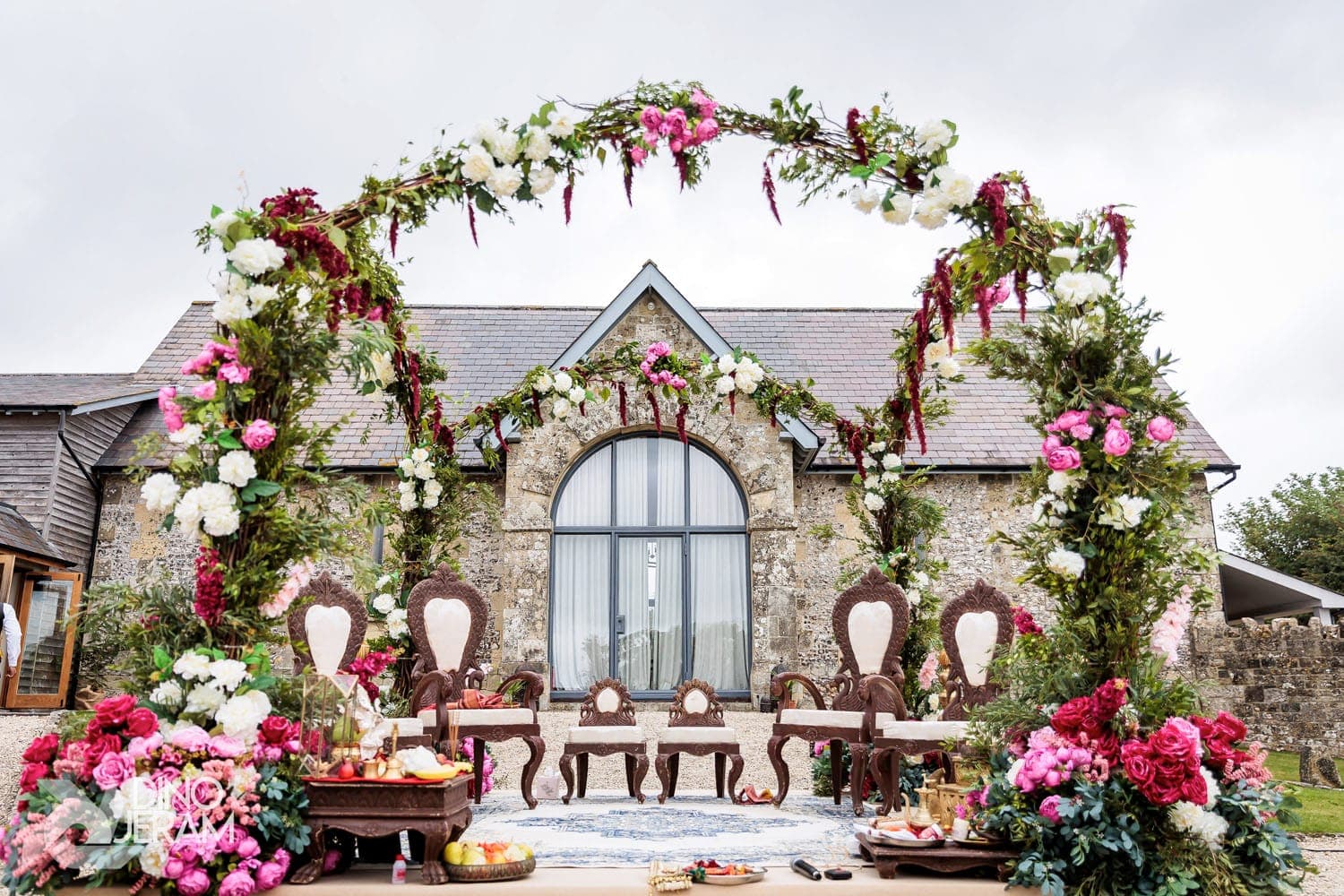 Wild floral ring with pink roses frames dark wooden chairs before a stone barn Wild floral ring with pink roses frames dark wooden chairs before a stone barn