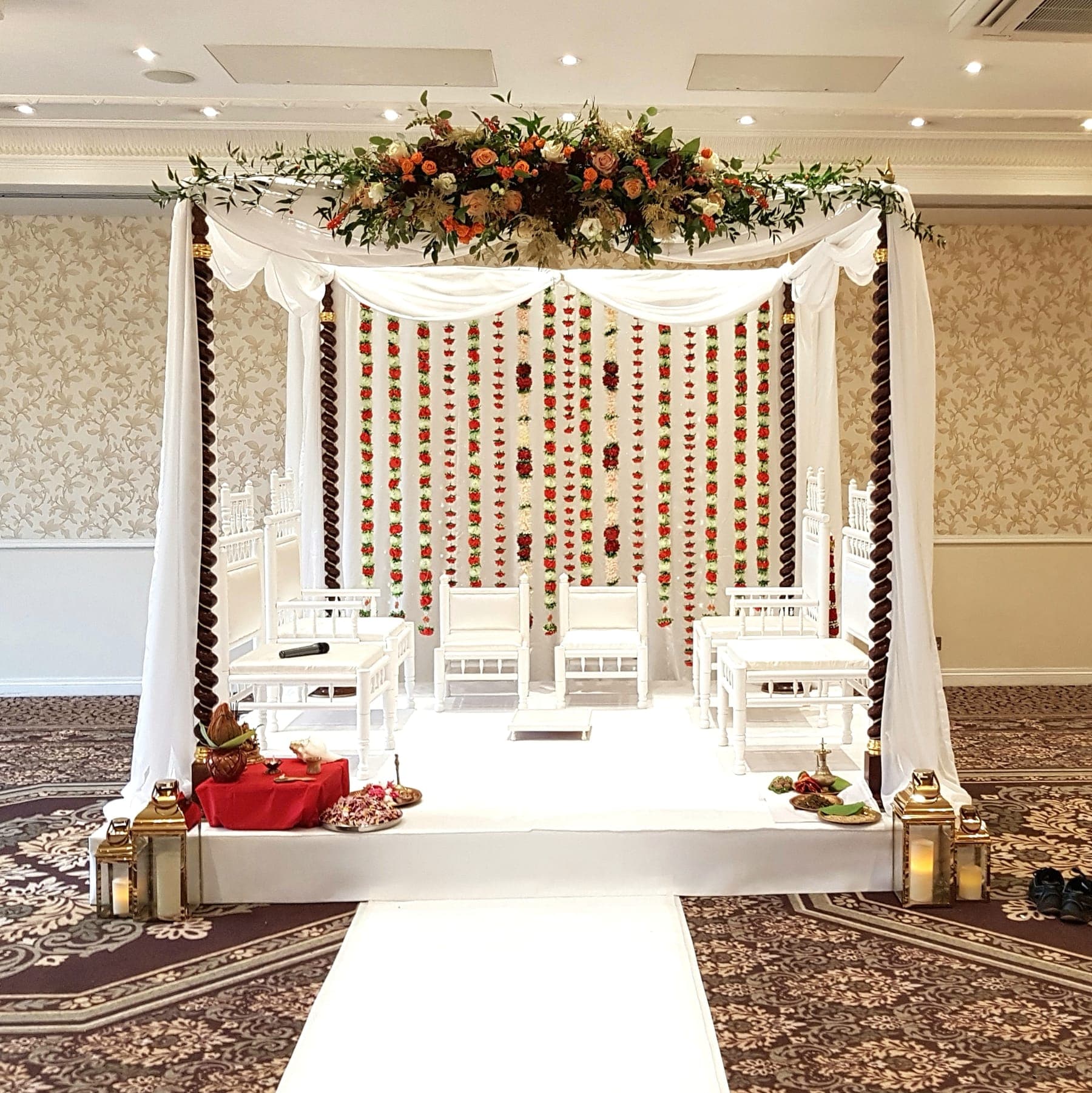 White frame with floral crown and red-orange garland curtain on a patterned carpet, lanterns at the base White frame with floral crown and red-orange garland curtain on a patterned carpet, lanterns at the base